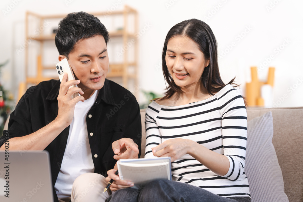 young Asian couple engaged in online shopping, wife woman holds notepad note list while husband man compares choices on smartphone. and call for price and discount, promotion. young Asian couple engaged in online shopping, wife woman holds notepad note list while husband man compares choices on smartphone. and call for price and discount, promotion.