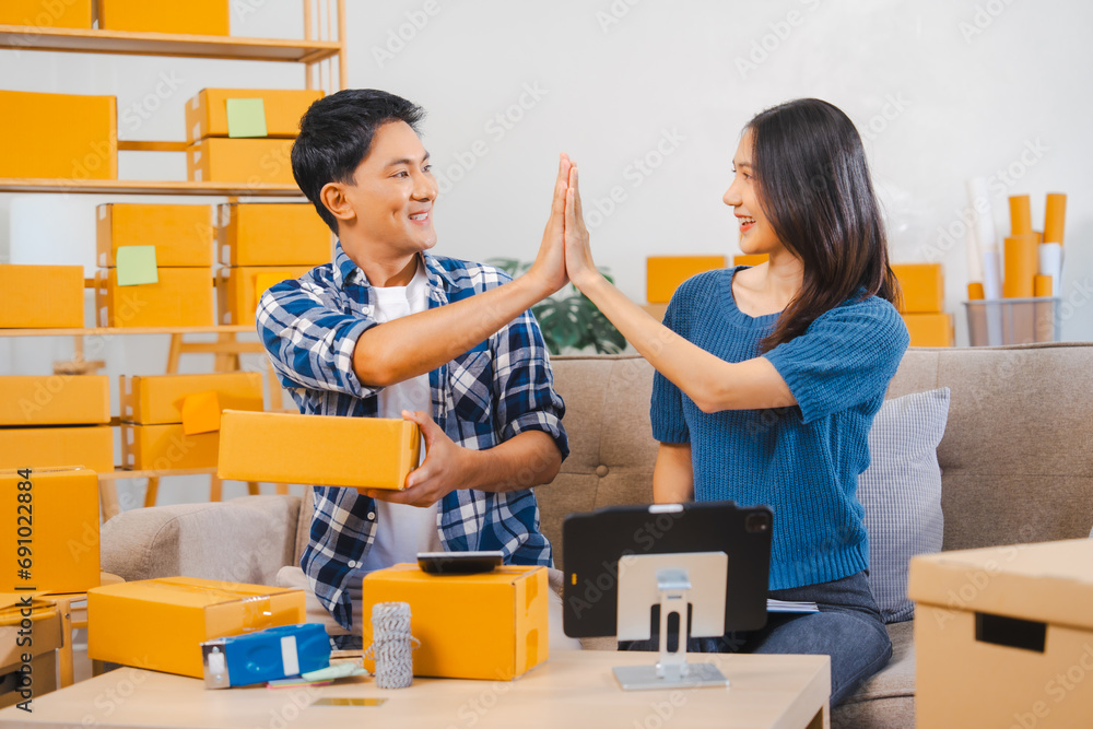 Asian people SME business woman and man couple joyfully showcasing products during a live stream in a room filled with packing boxes.