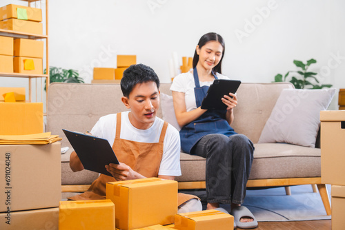 Asian man and woman in aprons work together, looking at paperwork and a laptop in a room filled with boxes.