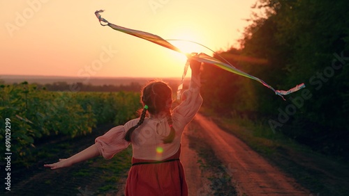 Happy girl runs along country road, plays with toy kite, sunset. Active Child runs with kite in sun. Kite hovers flies in hand of kid. Girl with toy kite. Child girl run play, dream of fly, traveling
