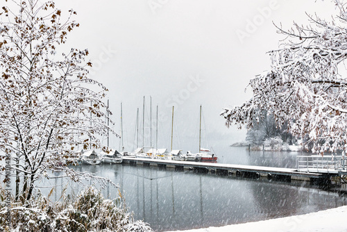 Photograph taken at Lake Caldonazzo in Trentino Alto Adige, Italy during a snowfall in November 2023. In the photo you can see some boats moored on a pier. photo