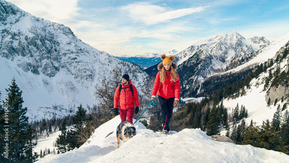 Active couple hiking on extreme terrain in the snowy mountains with their dog in winter - obrazy, fototapety, plakaty 