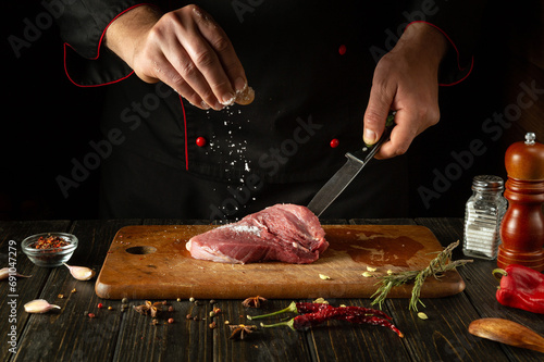 The cook sprinkles salt on the raw meat. Preparing beef steak before baking. Working environment in a restaurant or cafe kitchen photo