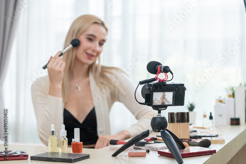 Young woman making beauty and cosmetic tutorial video content for social media. Beauty blogger smiles to camera while showing how to apply mascara to audience or followers. Blithe