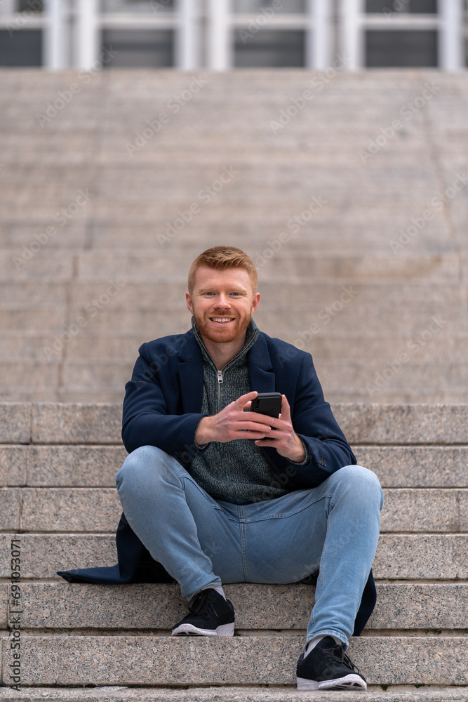 Red-haired adult seated on stairs, smiling at the camera while texting on a smartphone, effortlessly juggling tasks with a friendly demeanor Red-haired adult seated on stairs, smiling at the camera while texting on a smartphone, effortlessly juggling tasks with a friendly demeanor