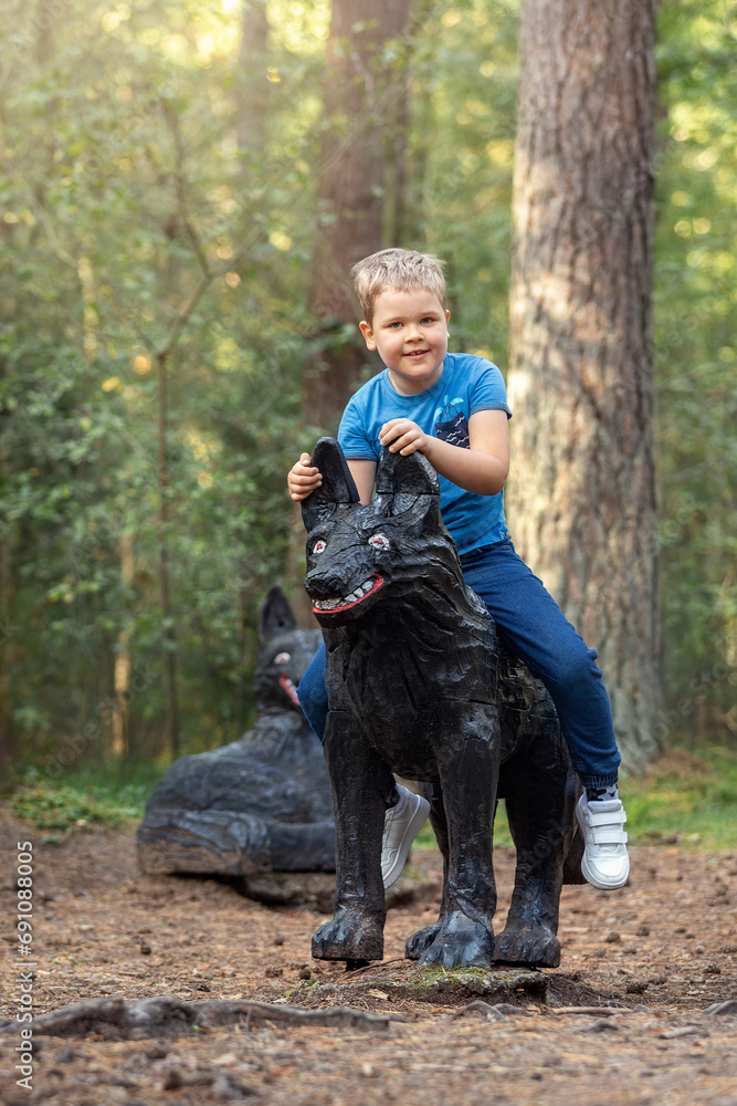 A cheerful smiling boy is riding on a black wolf sculpture on the background of a fairy forest. Vertical photo