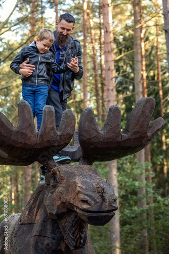 A father with his little boy on a large wooden elk antlers in the forest, tree tops in the background.