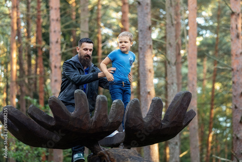 Low angle portrait in height father and son on large wooden elk antlers in the forest. A father holds his child firmly so that he does not fall photo