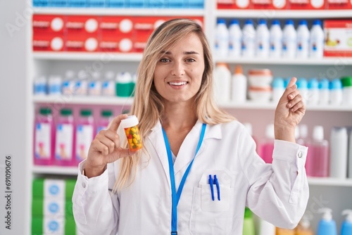 Young blonde woman working at pharmacy drugstore holding pills smiling happy pointing with hand and finger to the side