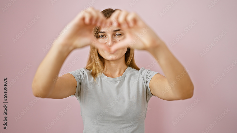 Cheerful young blonde woman confidently flashing a heart sign with hands, striking a casual pose over a pink isolated background. fashionable female enjoying her positive vibes & vibrant lifestyle.
