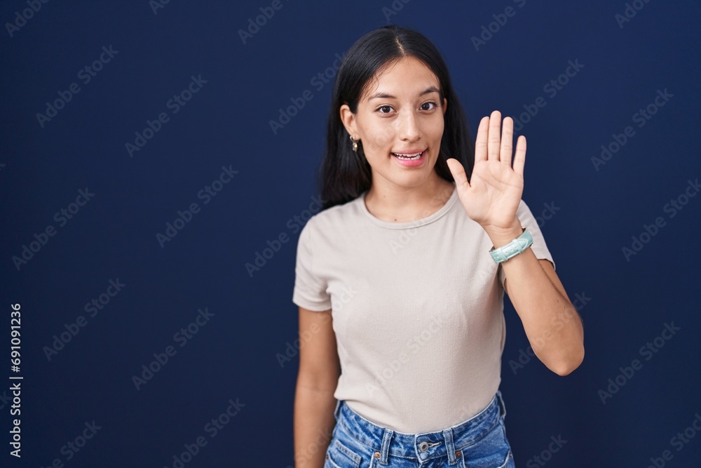 Young hispanic woman standing over blue background waiving saying hello happy and smiling, friendly welcome gesture Young hispanic woman standing over blue background waiving saying hello happy and smiling, friendly welcome gesture