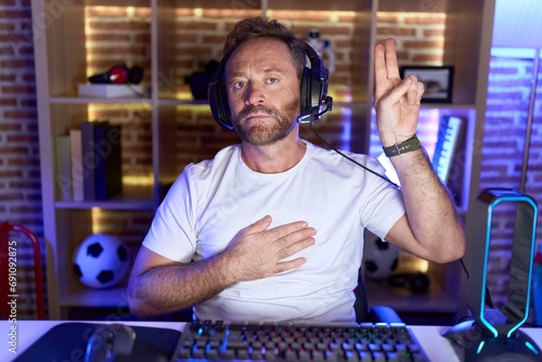 Middle age man with beard playing video games wearing headphones smiling swearing with hand on chest and fingers up, making a loyalty promise oath