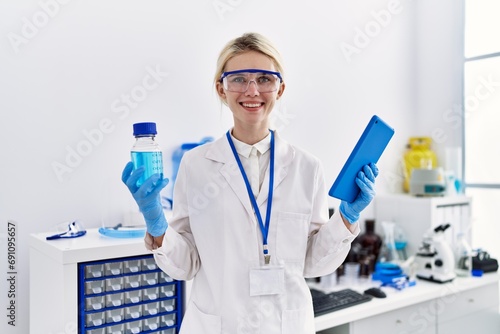 Young blonde woman scientist using touchpad holding bottle with liquid at laboratory