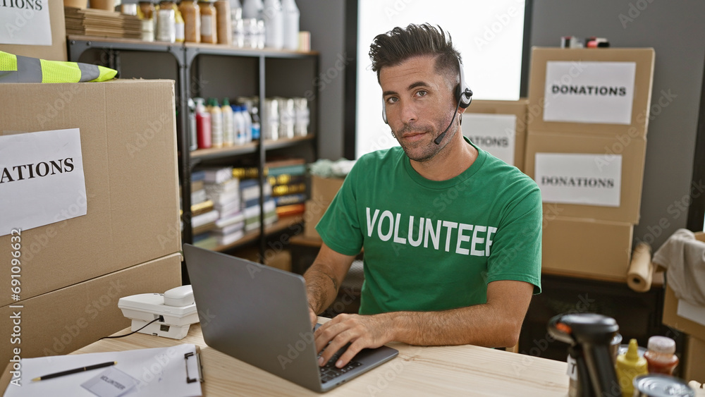 Handsome young hispanic man volunteering at community charity center, sitting at table, working online on laptop with headphones, offering social support with a warm smile. Handsome young hispanic man volunteering at community charity center, sitting at table, working online on laptop with headphones, offering social support with a warm smile.