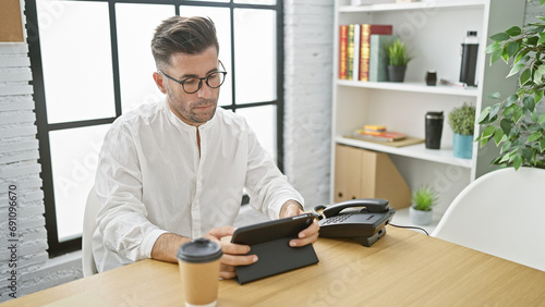 El jefe, portrait of a young, handsome hispanic man seriously working on his touchpad device at his office desk, embodying success in the digital era.