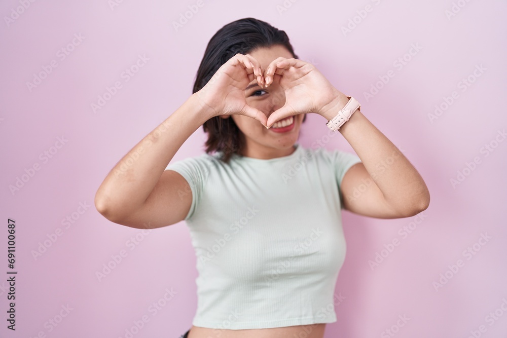 Hispanic young woman standing over pink background doing heart shape with hand and fingers smiling looking through sign Hispanic young woman standing over pink background doing heart shape with hand and fingers smiling looking through sign