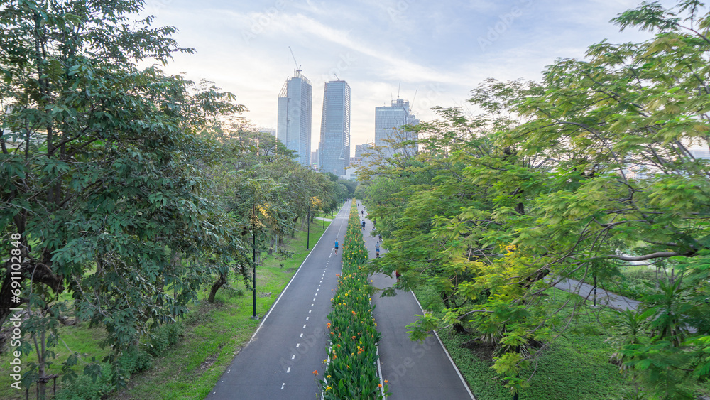 A picture of a public park in the middle of a large city. with dense high-rise buildings There were still people out jogging in the garden. Amidst the shady atmosphere of various types of trees.