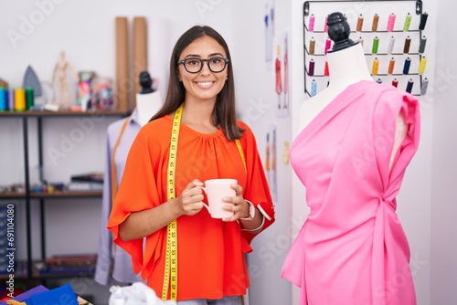 Young beautiful hispanic woman tailor drinking coffee standing by manikin at tailor shop
