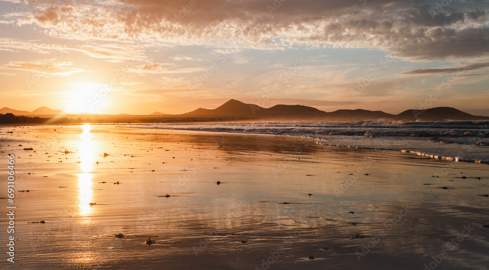 Panoramic view of beautiful golden hour on the Famara. Beach in Lanzarote - Canary Islands.  Photo of sunset on the beach with reflection of ocean and big waves in ocean and dramatic cloudy sky.