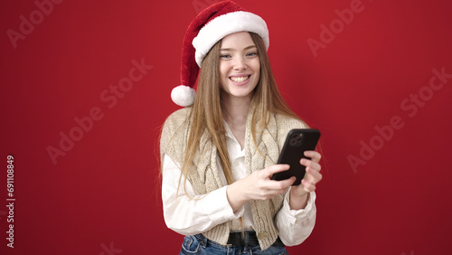 Young blonde woman wearing christmas hat using smartphone over isolated red background