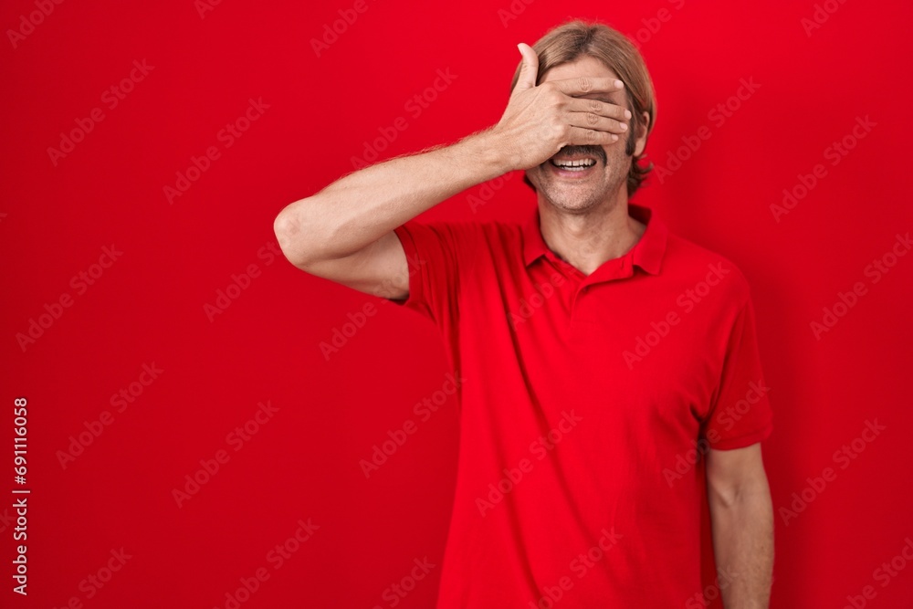Caucasian man with mustache standing over red background smiling and laughing with hand on face covering eyes for surprise. blind concept. Caucasian man with mustache standing over red background smiling and laughing with hand on face covering eyes for surprise. blind concept.