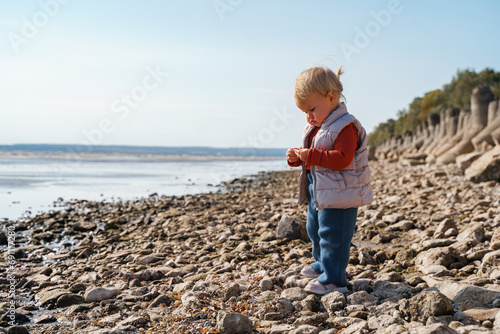 Cute little girl kid toddler in a terracotta turtleneck on a stone beach by the sea, cold season, Caucasian blonde child alone