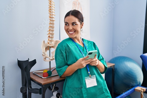 Young beautiful hispanic woman physiotherapist smiling confident using smartphone at rehab clinic