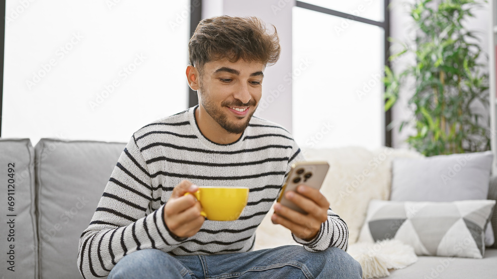 Young, happy arab man relaxing at home, enjoying his coffee while texting on his smartphone. smiling confidently, this handsome adult gives off a serene expression. Young, happy arab man relaxing at home, enjoying his coffee while texting on his smartphone. smiling confidently, this handsome adult gives off a serene expression.