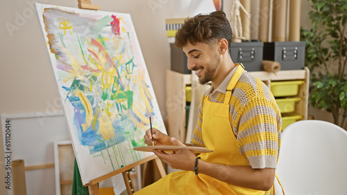 Confident young arab art student smiling as he expertly draws in a vibrant university art studio, harnessing his creativity with brush and palette
