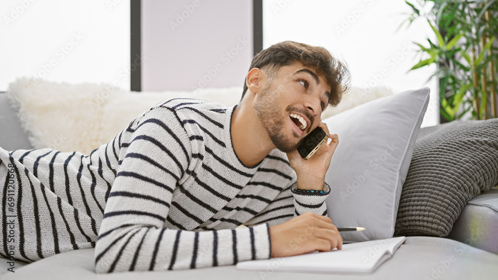 Engrossed in literature, young arabian man smiling confidently while speaking on phone and writing in notebook on sofa at home