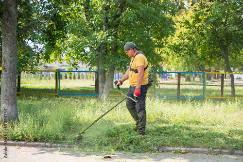 Man gardener mowing ragweed bushes, ambrosia artemisiifolia that causing allergy summer and autumn with electric or petrol lawn trimmer along the city street. Man mows grass with a gasoline scythe.