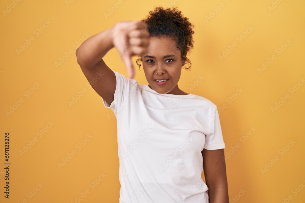 Young hispanic woman with curly hair standing over yellow background looking unhappy and angry showing rejection and negative with thumbs down gesture. bad expression. Young hispanic woman with curly hair standing over yellow background looking unhappy and angry showing rejection and negative with thumbs down gesture. bad expression.