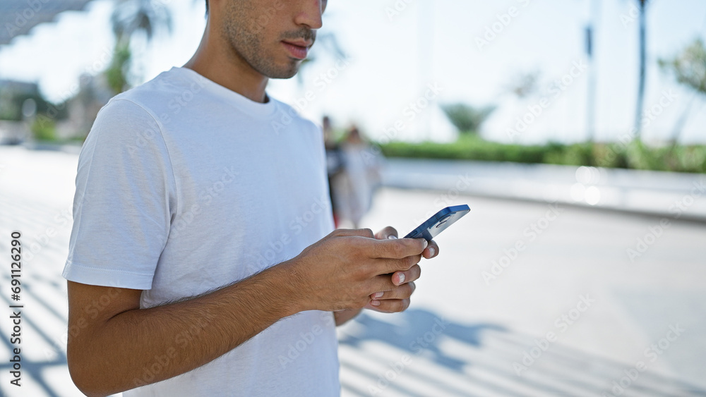 Cool looking young hispanic man, with serious expression, deeply engrossed in texting on smartphone at park, emanating attractive relaxed lifestyle amidst green nature under sunny outdoors. Cool looking young hispanic man, with serious expression, deeply engrossed in texting on smartphone at park, emanating attractive relaxed lifestyle amidst green nature under sunny outdoors.