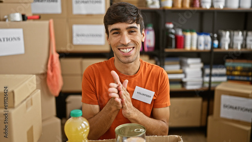 Young and handsome hispanic man, wholeheartedly clapping in applause, sitting at a table serving as a confident volunteer in support of the charity center, celebrating the altruism indoors photo