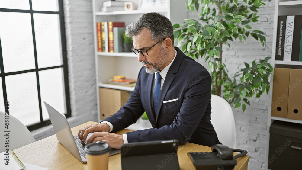Attractive, grey-haired young hispanic man, an elegant professional, concentrating hard in the office. he's working seriously, managing business success from his laptop at his desk. Attractive, grey-haired young hispanic man, an elegant professional, concentrating hard in the office. he's working seriously, managing business success from his laptop at his desk.