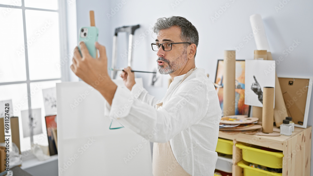 Handsome young hispanic, grey-haired male artist engrossed in a serious video call at indoor art studio, painting with paintbrush on canvas, learning and discussing art lessons. Handsome young hispanic, grey-haired male artist engrossed in a serious video call at indoor art studio, painting with paintbrush on canvas, learning and discussing art lessons.