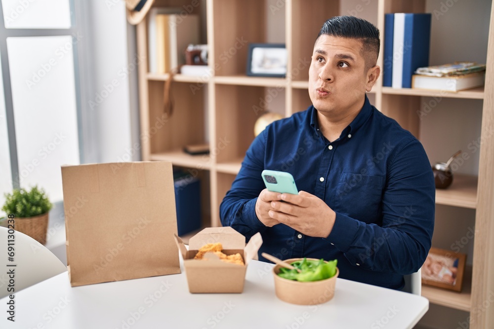 Hispanic young man eating take away food using smartphone smiling looking to the side and staring away thinking. Hispanic young man eating take away food using smartphone smiling looking to the side and staring away thinking.