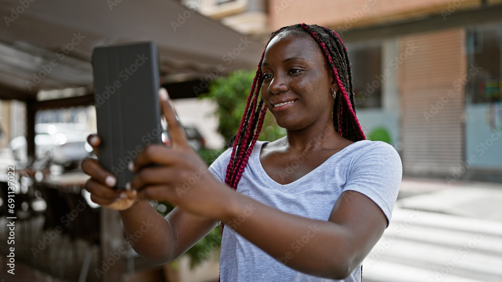 Confident african american woman basking in sunlight, making selfie with touchpad, smiling joyously at urban coffee shop terrace