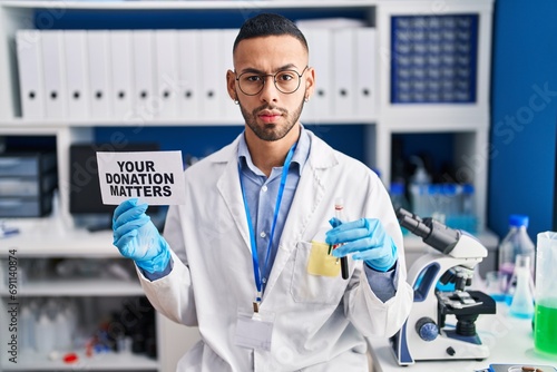 Young hispanic man working at scientist laboratory holding your donation matters holding blood sample skeptic and nervous  frowning upset because of problem. negative person.