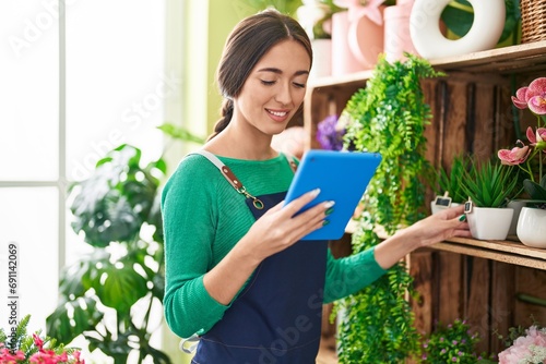 Young beautiful hispanic woman florist using touchpad holding plant of shelving at flower shop