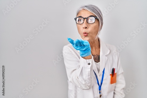 Middle age woman with grey hair wearing scientist robe looking at the camera blowing a kiss with hand on air being lovely and sexy. love expression.