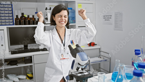 Celebrating breakthrough  young and beautiful hispanic woman scientist  joyfully working with a microscope in the lab  makes a momentous discovery