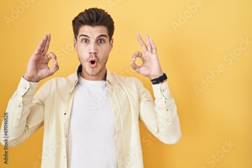 Young hispanic man standing over yellow background looking surprised and shocked doing ok approval symbol with fingers. crazy expression