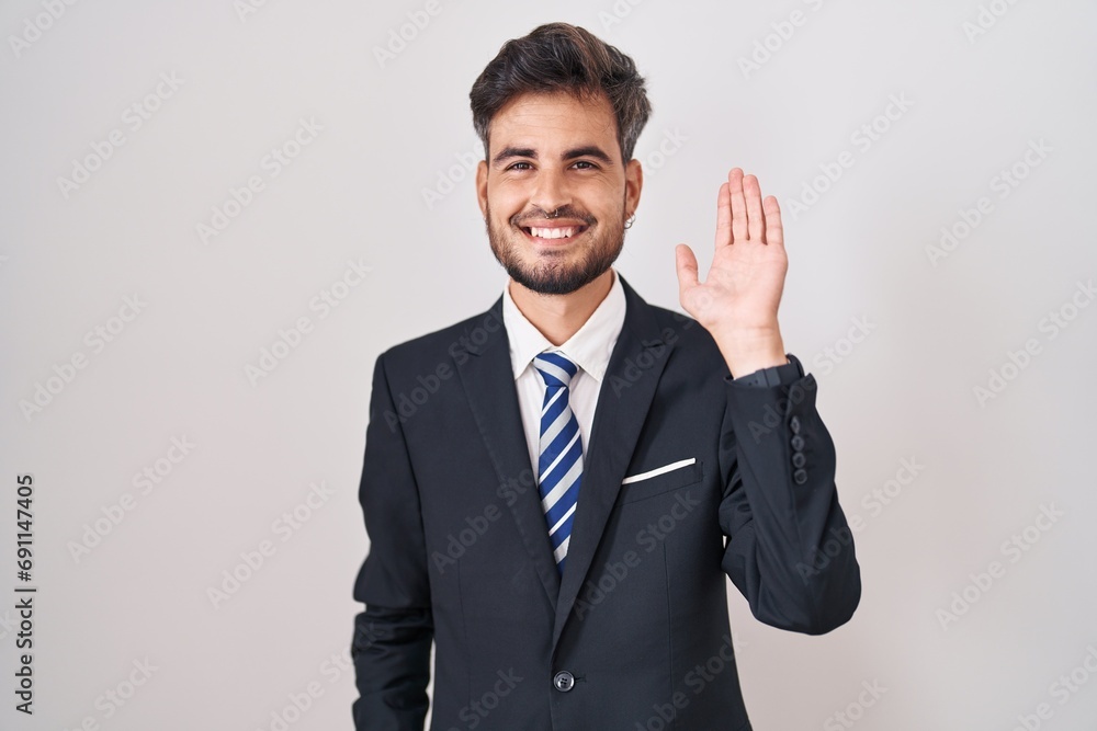 Young hispanic man with tattoos wearing business suit and tie waiving saying hello happy and smiling, friendly welcome gesture Young hispanic man with tattoos wearing business suit and tie waiving saying hello happy and smiling, friendly welcome gesture