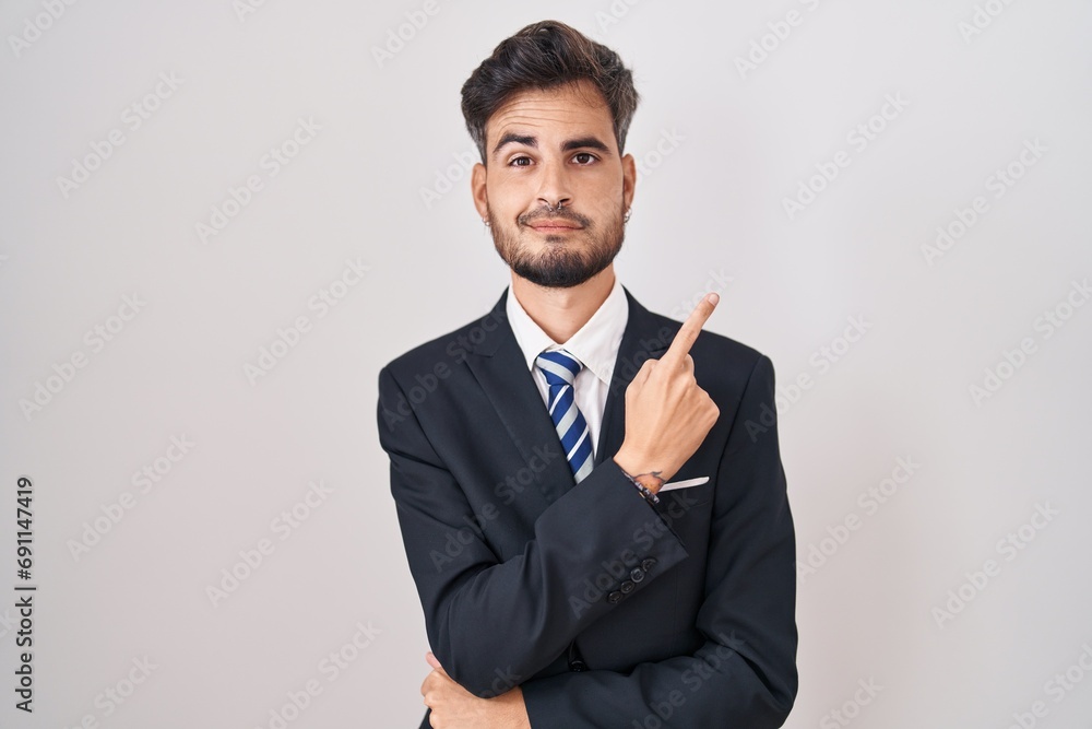 Young hispanic man with tattoos wearing business suit and tie pointing with hand finger to the side showing advertisement, serious and calm face Young hispanic man with tattoos wearing business suit and tie pointing with hand finger to the side showing advertisement, serious and calm face