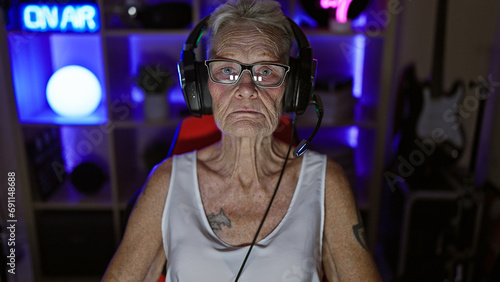 Serious grey-haired senior woman streamer engrossed in a nighttime digital game at home, relaxed yet focused, sitting with headset and gamepad in her gaming room photo