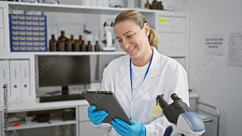 Blonde woman scientist's jubilant smile at the lab, young and confident, enjoying her touchpad while engaging with innovative medical technology
