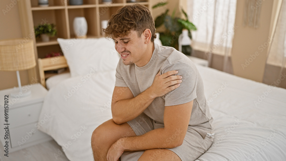 Worried young hispanic man, in pyjamas, suffering intense shoulder pain. unhappy, he lies awake in his home, touching his injured arm on his bed in the morning.