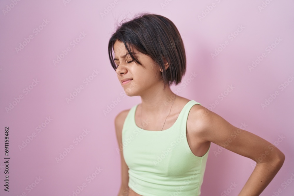Young girl standing over pink background suffering of backache, touching back with hand, muscular pain Young girl standing over pink background suffering of backache, touching back with hand, muscular pain