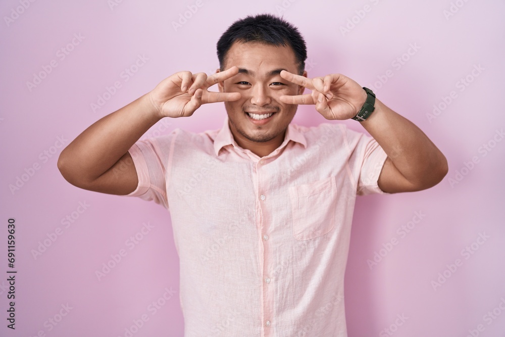 Chinese young man standing over pink background doing peace symbol with fingers over face, smiling cheerful showing victory Chinese young man standing over pink background doing peace symbol with fingers over face, smiling cheerful showing victory